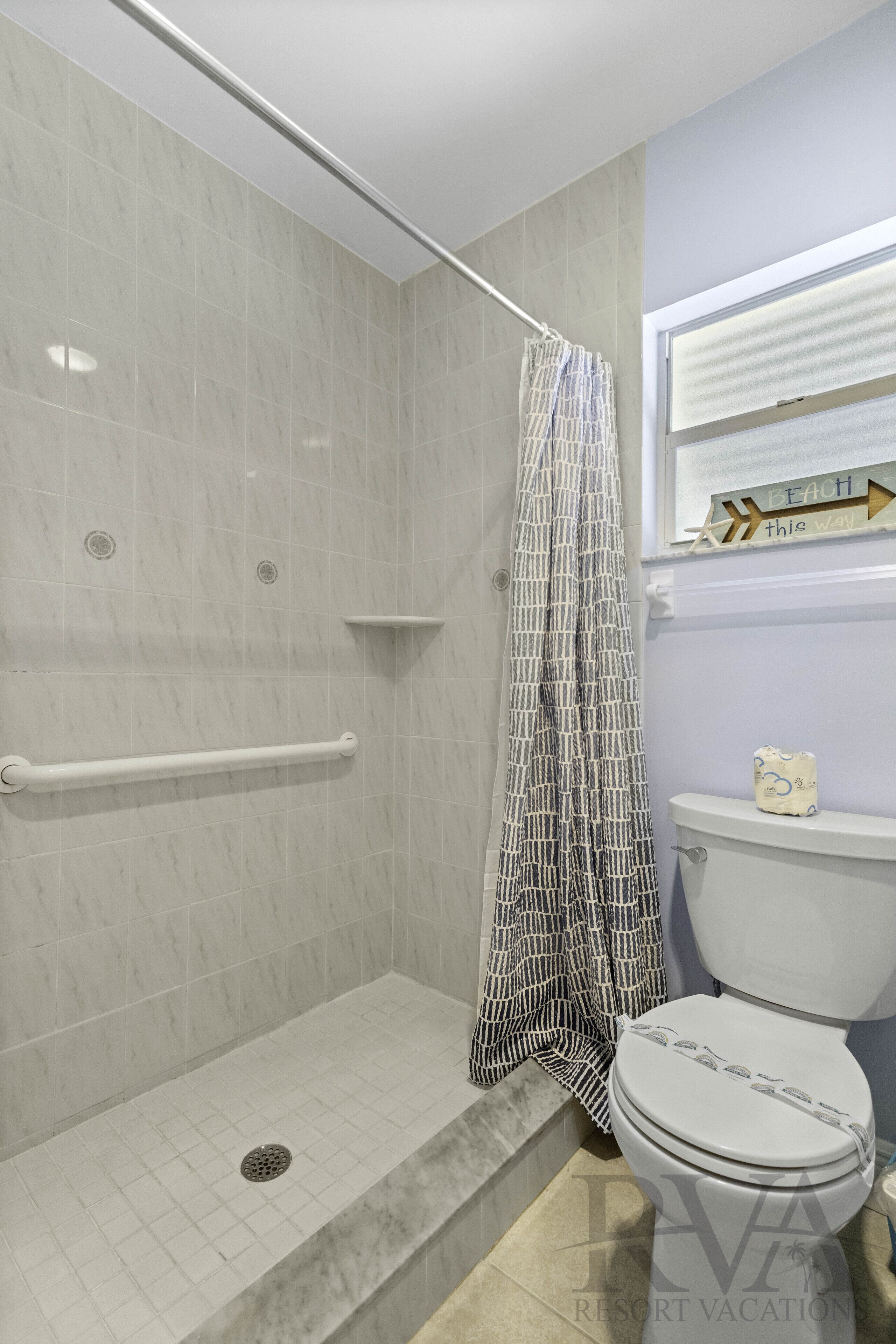 A bathroom with a shower, a toilet, and a window with a beach sign.