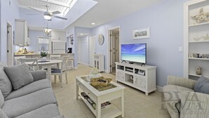 A modern living room with a flat-screen TV, a ceiling fan, and a dining area with a kitchen in the background. - Gulf Getaway – Beach Castle #12 by RVA (Longboat Key)