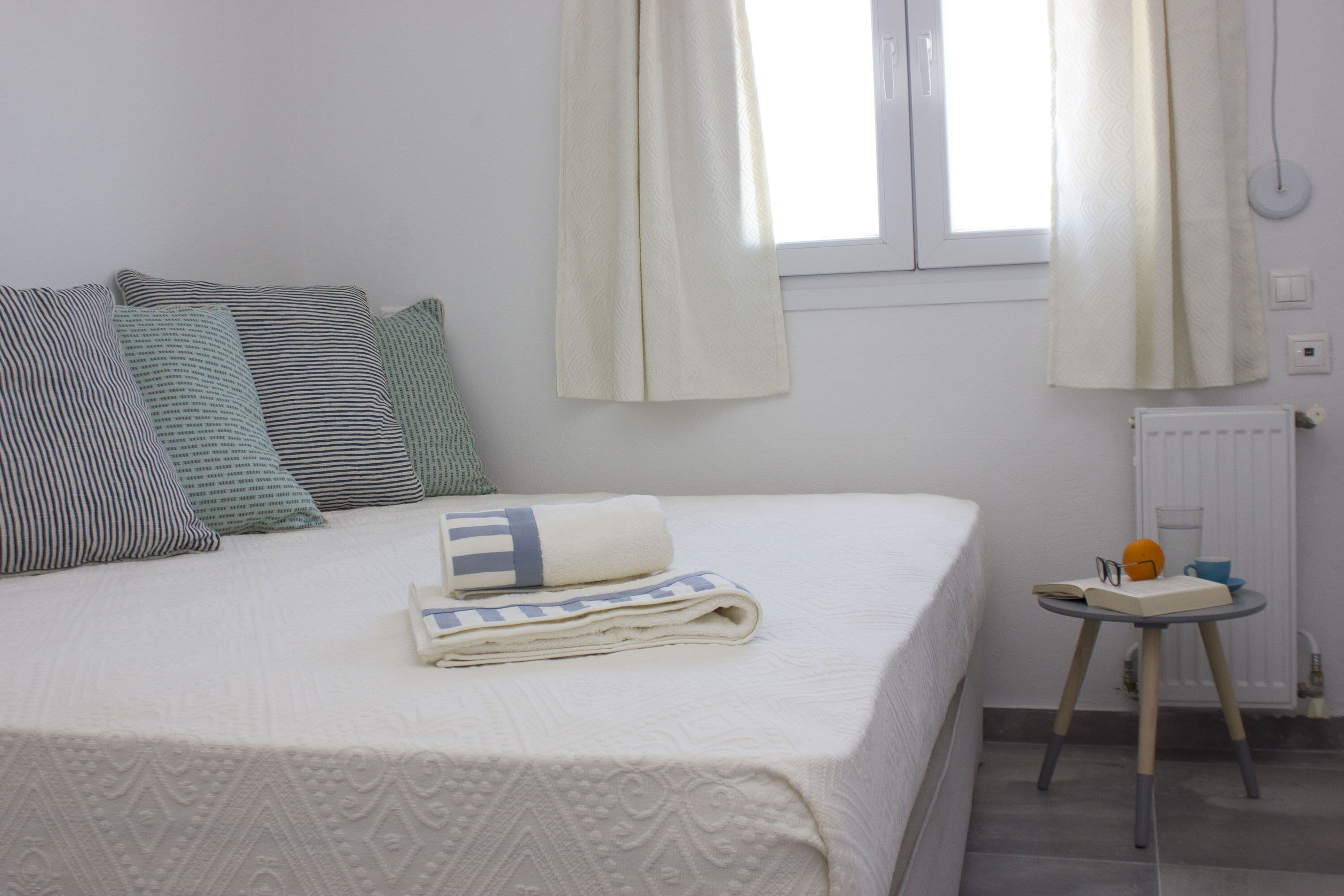 A neatly made bed with a folded towel, pillows, and a small bedside table with a book, glasses, and a cup.