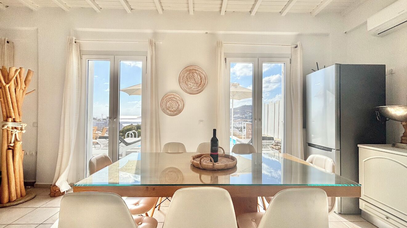 A dining area with a glass table, white chairs, and a view of the sea through large windows.