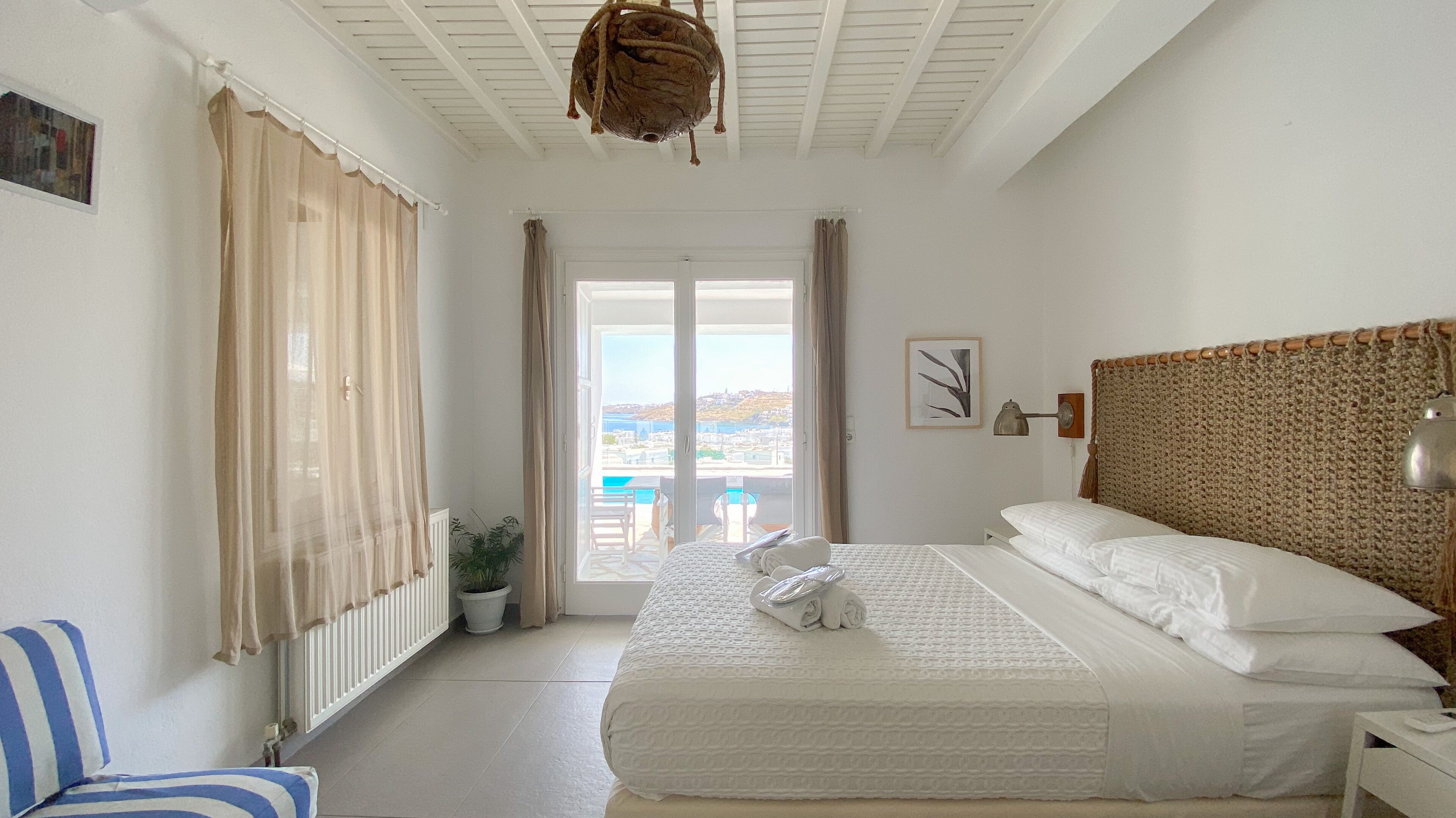 A bedroom with a bed, a nightstand, a lamp, a view of the sea, and a potted plant.