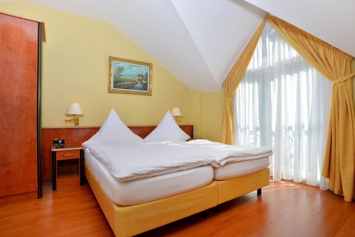 A hotel room with a bed, two bedside lamps, a painting on the wall, and a window with curtains.