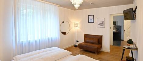 A hotel room with a large bed, a brown armchair, a small table with a vase, and a television on the wall.