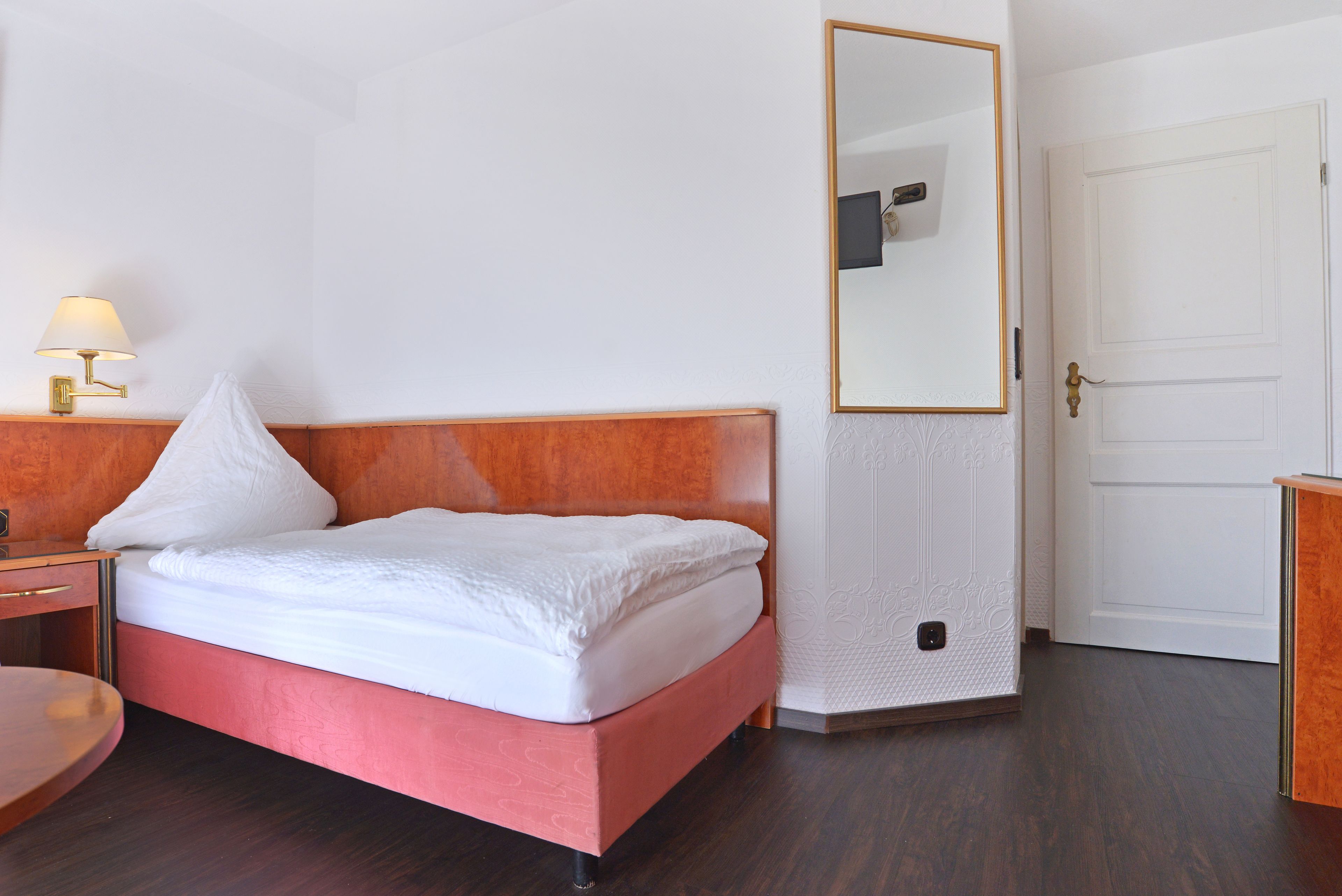 A neatly made bed with a wooden headboard, a bedside table with a lamp, and a large mirror on the wall.