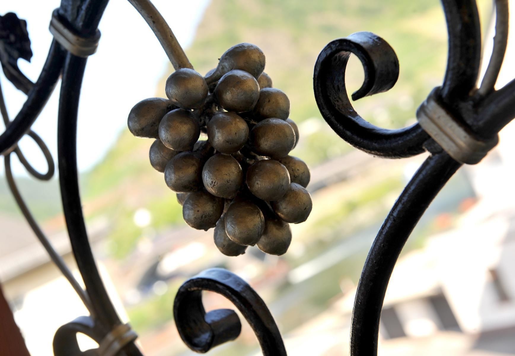 Une balustrade de balcon en fer forgé, ornée d’une grappe de raisins suspendue.