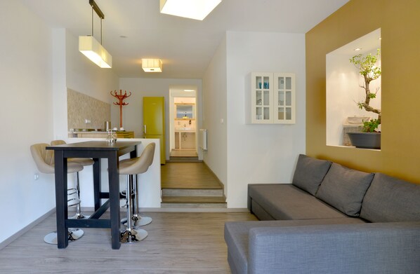 A modern living room with a grey sofa, a dining area with a table and chairs, and a staircase leading to another room.