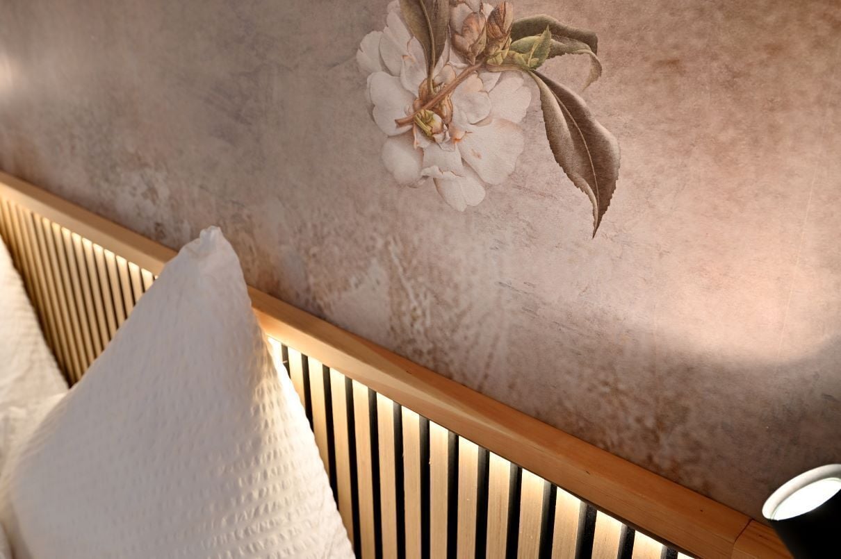 Una cama con cabecera de madera, almohada blanca y pared con estampado floral.