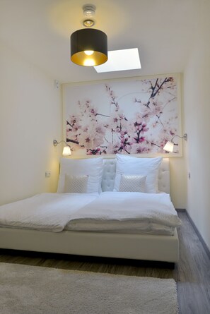 A neatly made bed with white linens and a fluffy white rug, a wall-mounted light fixture, and a large floral artwork above the bed. - Hotel Zehnthof (Cochem)