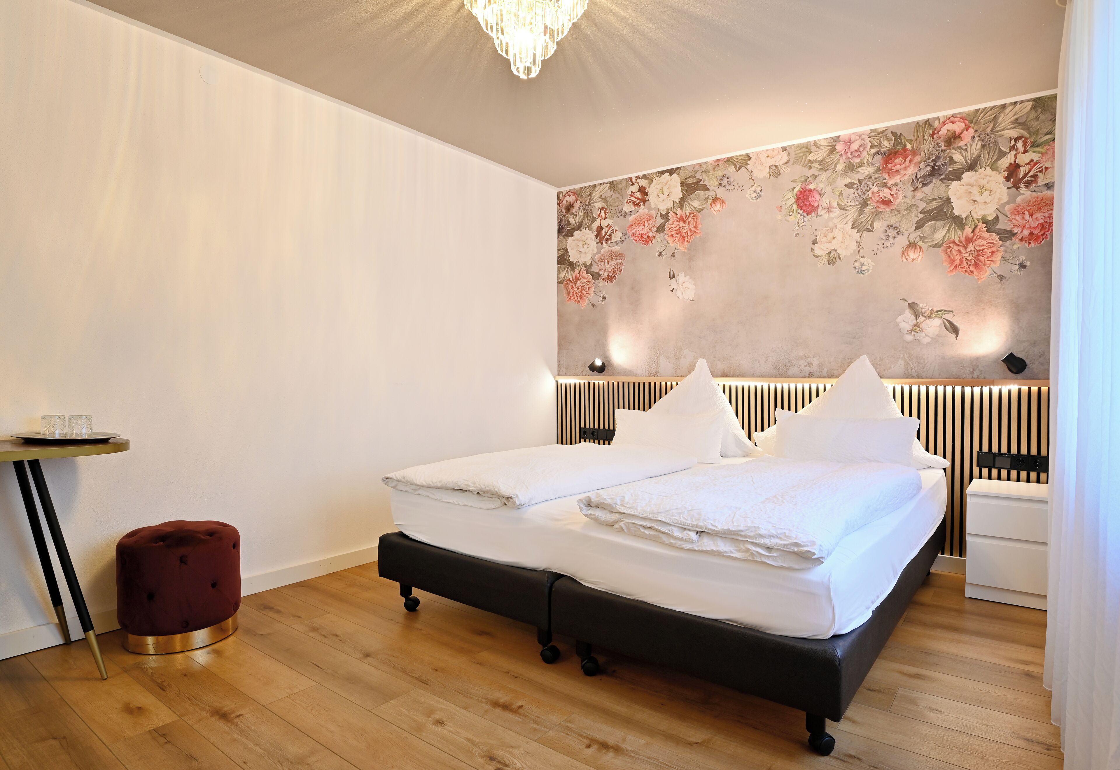 A modern bedroom with a double bed, a small red ottoman, a bedside table, and a floral wall mural.