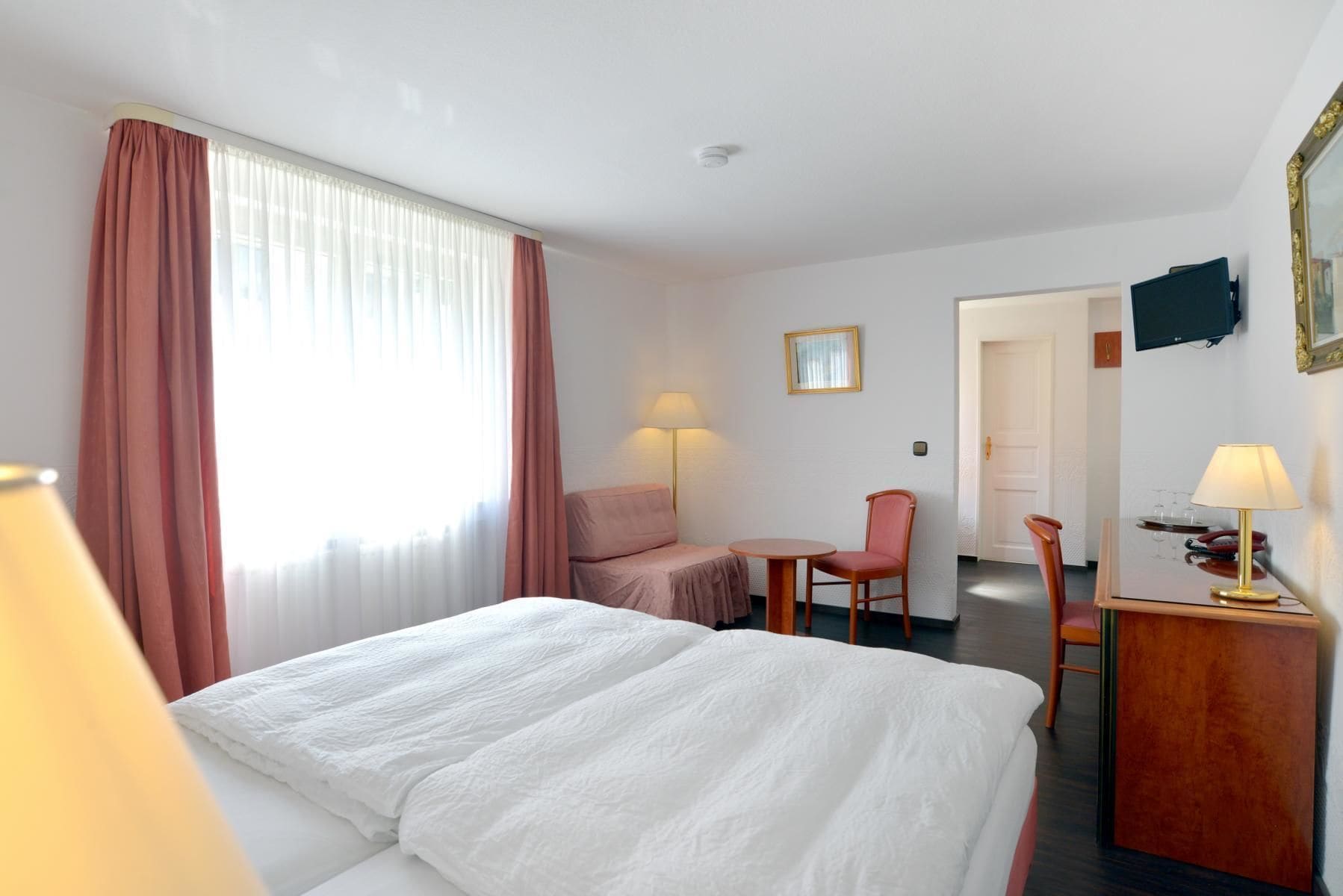 A hotel room with a bed, a desk, a chair, a lamp, and a television on the wall.