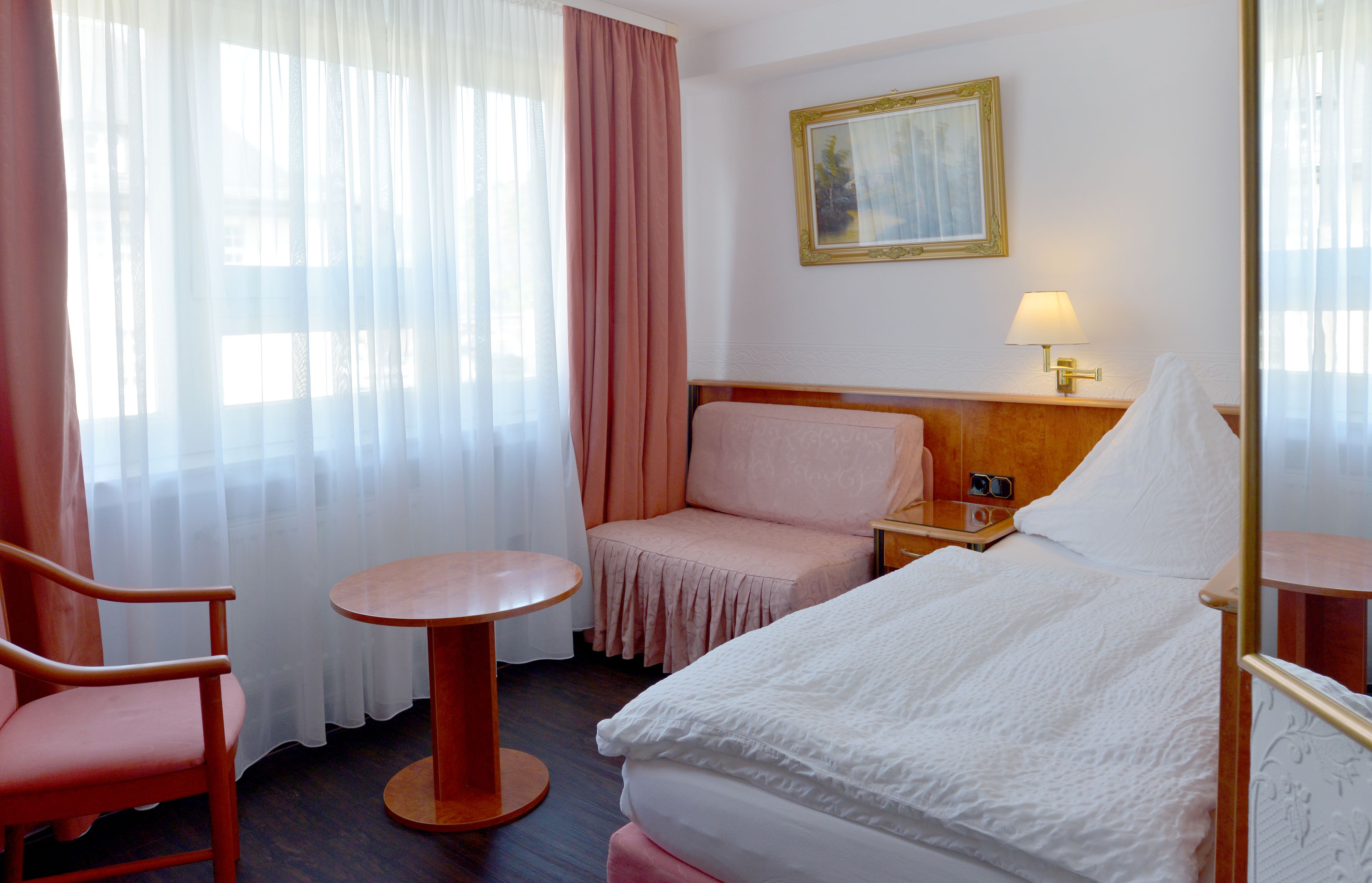 A hotel room with a bed, a chair, a small table, a nightstand, a lamp, and a painting on the wall.