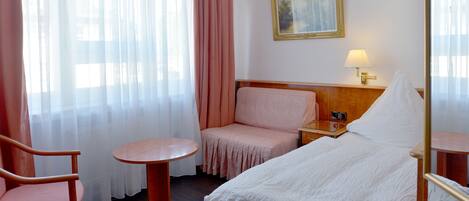Habitación de hotel con cama, una silla, una mesita, una mesita de noche, una lámpara y un cuadro en la pared.