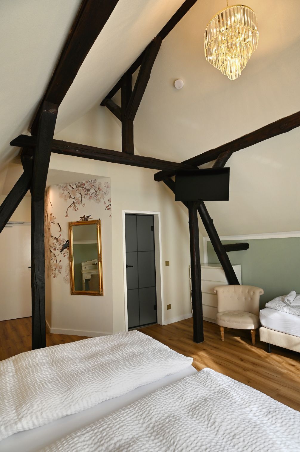 Une chambre avec des poutres apparentes, un lustre, un lit, une chaise et un miroir.