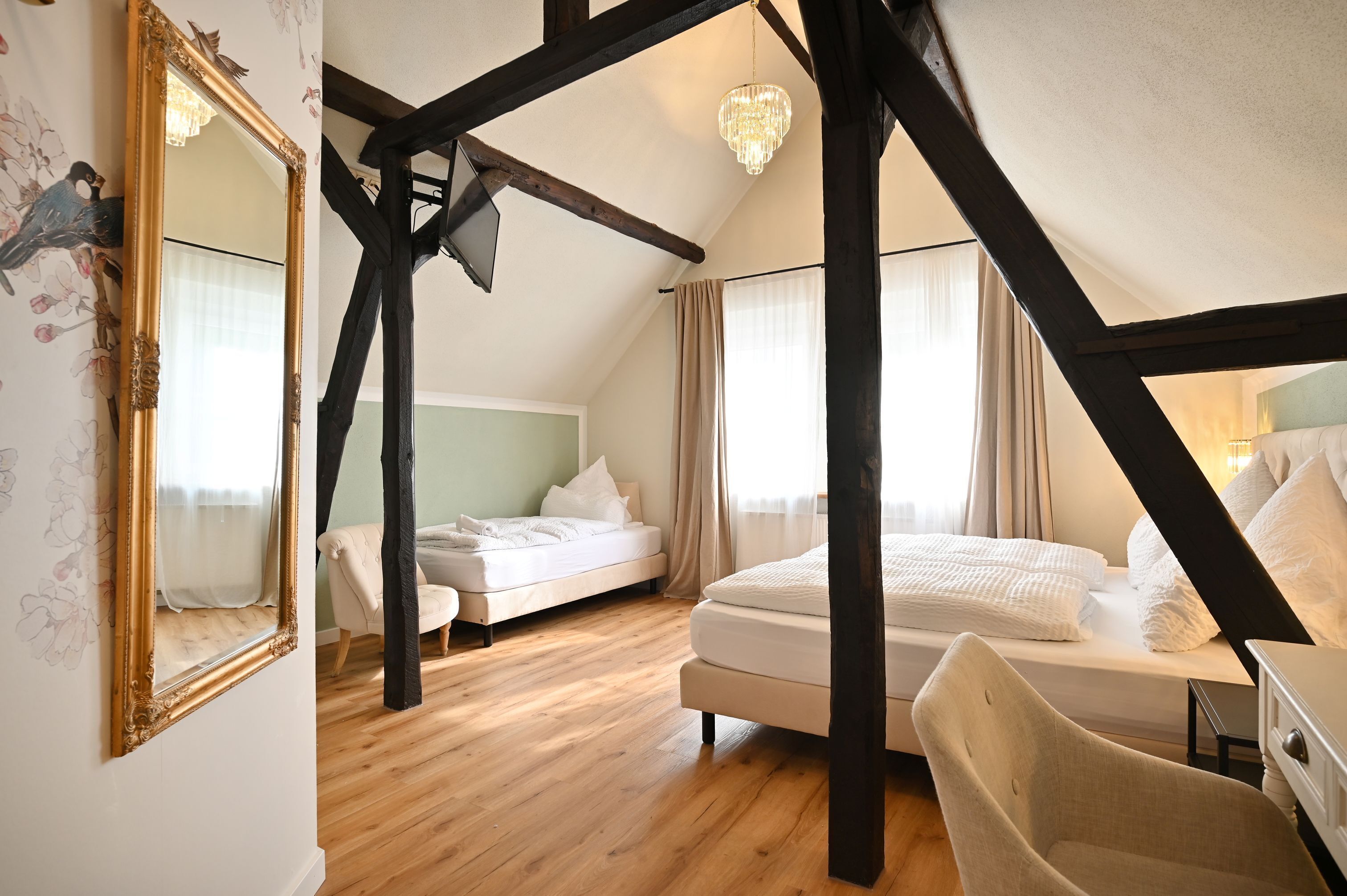 A bedroom with a sloped ceiling, wooden beams, a large mirror, two beds, and a chandelier.