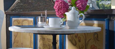 Una mesa con una jarrón blanco y flores rosadas, una silla blanca y una vista de un castillo en una colina.