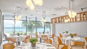 Een restaurant met ronde tafels gedekt voor diners, grote ramen met uitzicht op het buitengebied, en hangende pendantlampen.