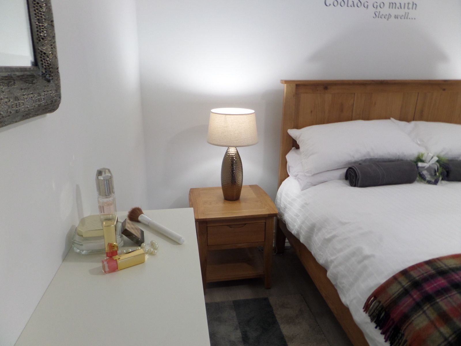 A neatly made bed with a plaid blanket, a wooden nightstand with a lamp, and a wall with a motivational quote.