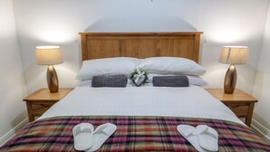 A neatly made bed with white linens, two pillows, a plaid blanket, and a pair of slippers on the bed.