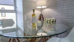 A glass-top dining table with white chairs, a bottle of wine, and a lamp on a brick wall backdrop.
