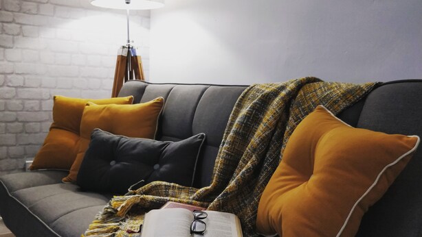A grey sofa with mustard and black cushions, a plaid throw blanket, and an open book with glasses on it.