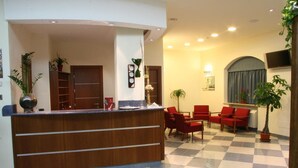 Zona de recepción de un hotel con piso a cuadros, un mostrador de madera, sillas rojas y plantas en macetas.