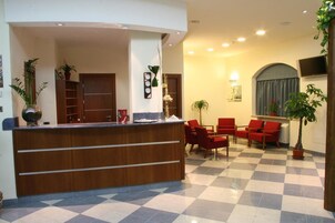 A hotel reception area with a checkered floor, a wooden reception desk, red chairs, and potted plants.
