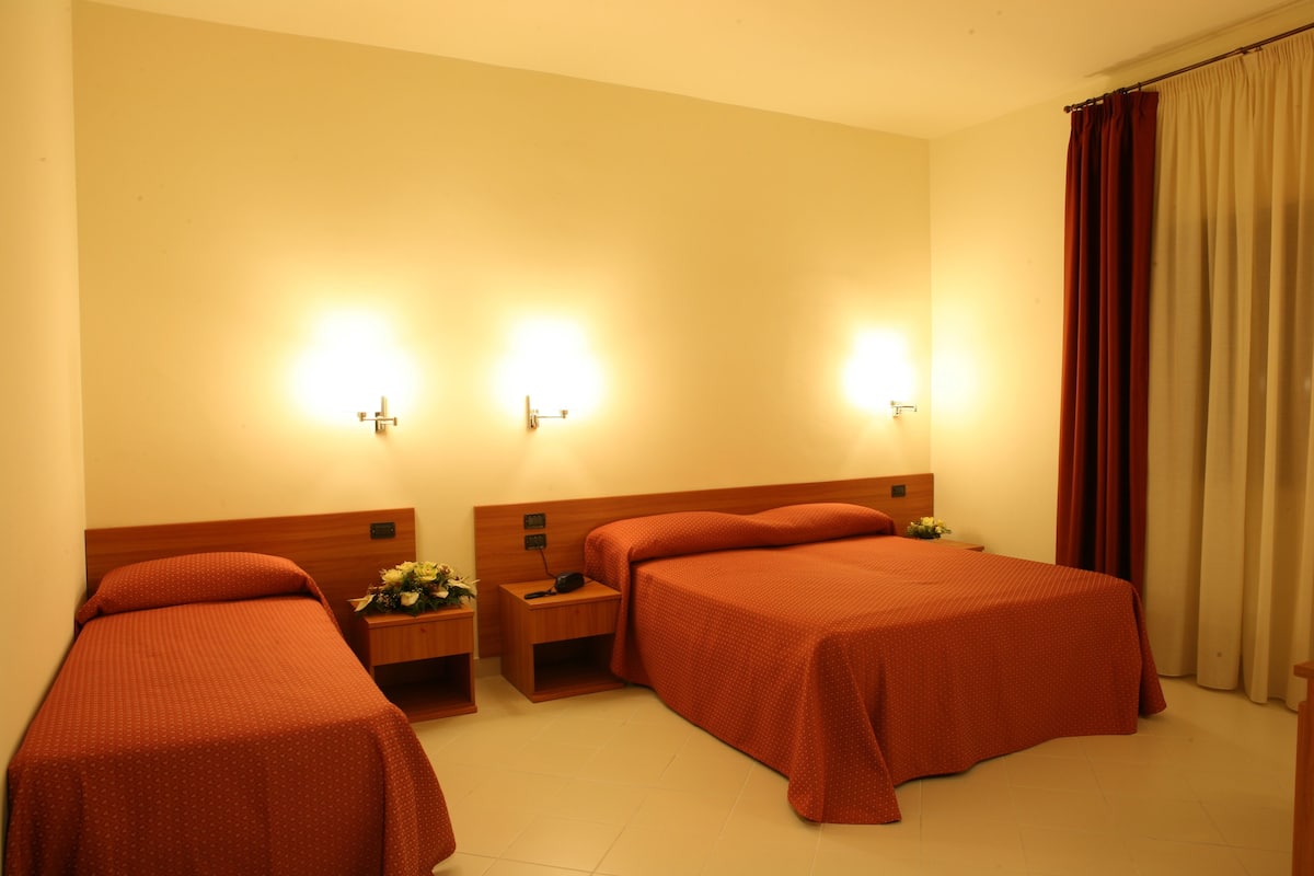 A hotel room with two single beds, a wooden headboard, bedside tables with lamps, and a curtain on the window.