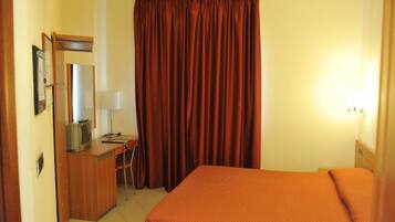 Habitació d'hotel amb un llit, una taula de treball amb una cadira, una televisió i una finestra gran amb cortines.