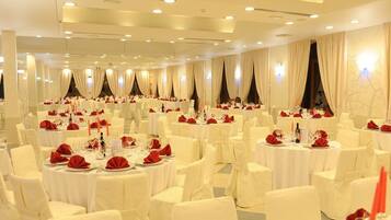 Un salón de banquetes con mesas redondas dispuestas para comer, sillas blancas y servilletas rojas.