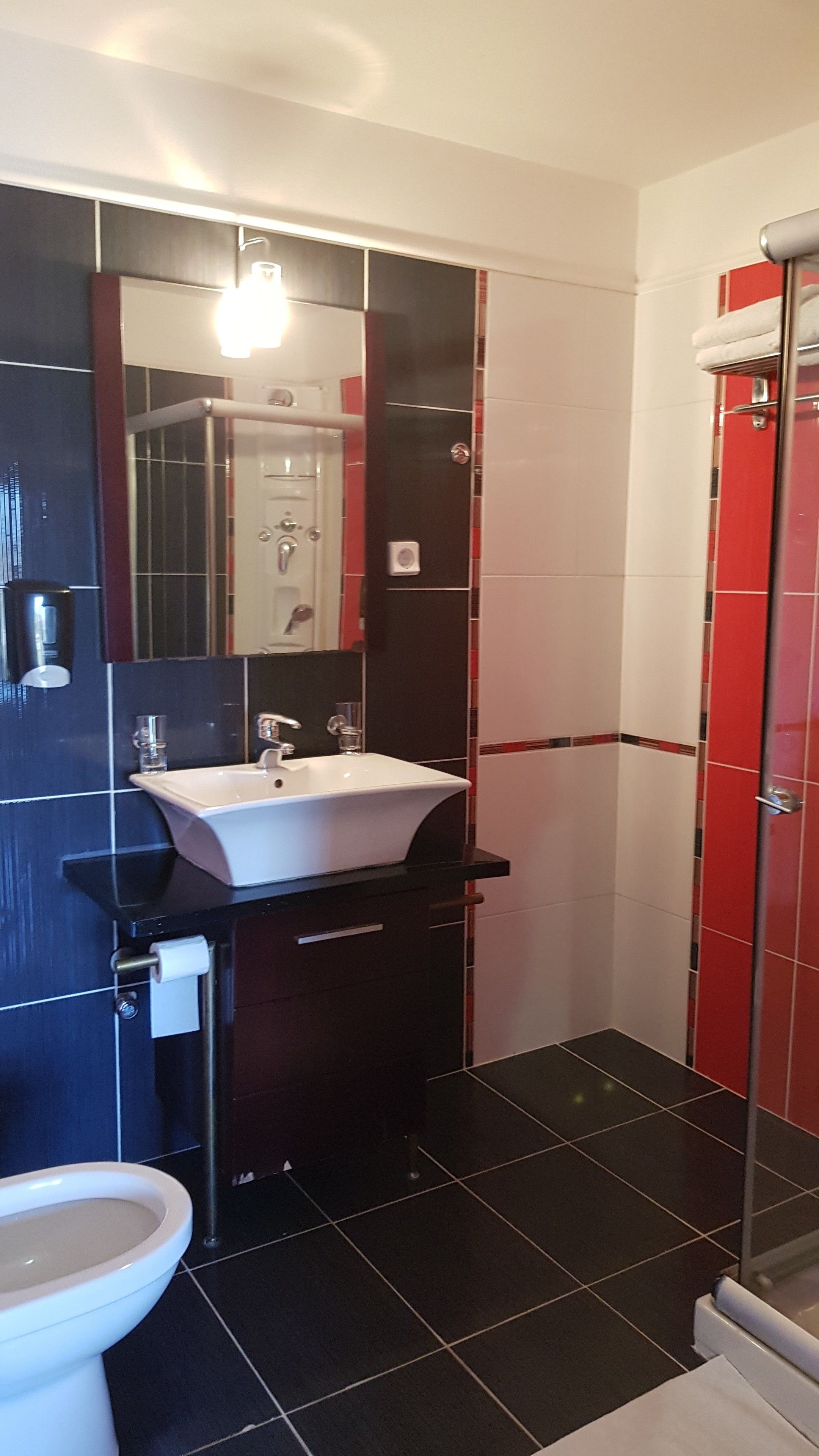 A bathroom with a white sink, black and red tiled walls, a glass shower enclosure, and a toilet.
