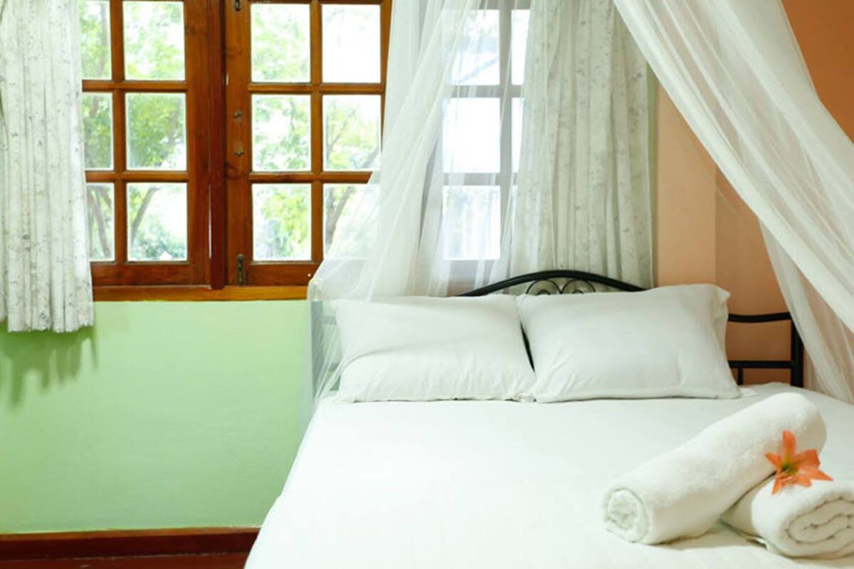 Ein Bett mit weißer Bettwäsche und ein zusammengerolltes Handtuch mit einer Blume, ein Fenster mit Vorhängen und eine grüne Wand.