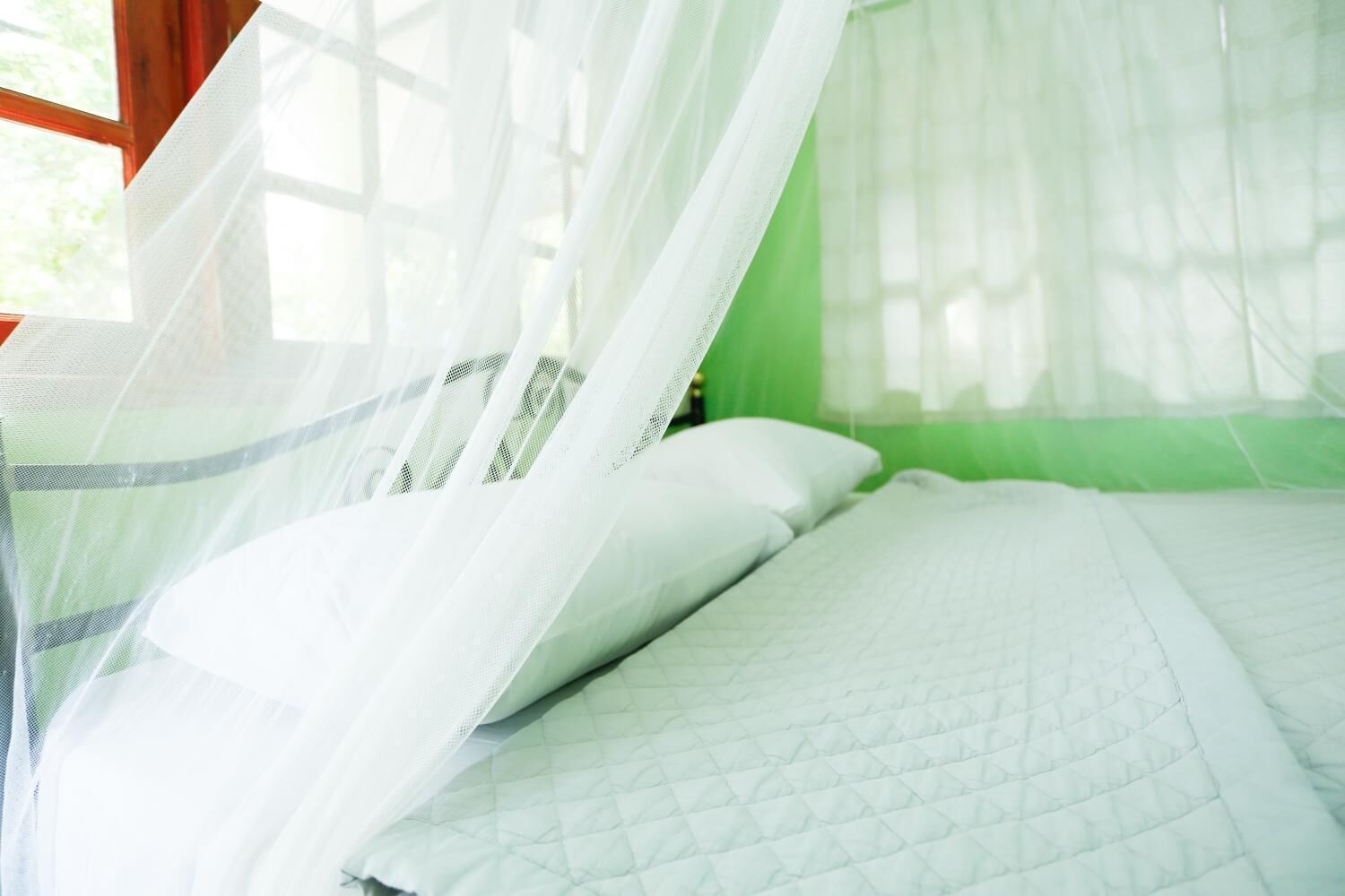 Una cama con una colcha blanca acolchada y almohadas blancas, bajo una mosquitera blanca.