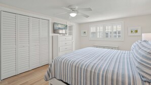 A bedroom with a bed, a ceiling fan, a dresser, a window with blinds, and a door with louvered shutters.