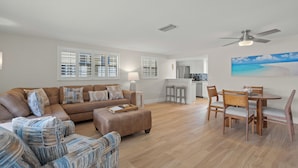 A spacious living room with a sofa, ottoman, and chairs, a dining area with a table and chairs, and a kitchen in the background.