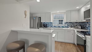A modern kitchen with white cabinetry, a stainless steel refrigerator, a tiled backsplash, and a window with blinds.