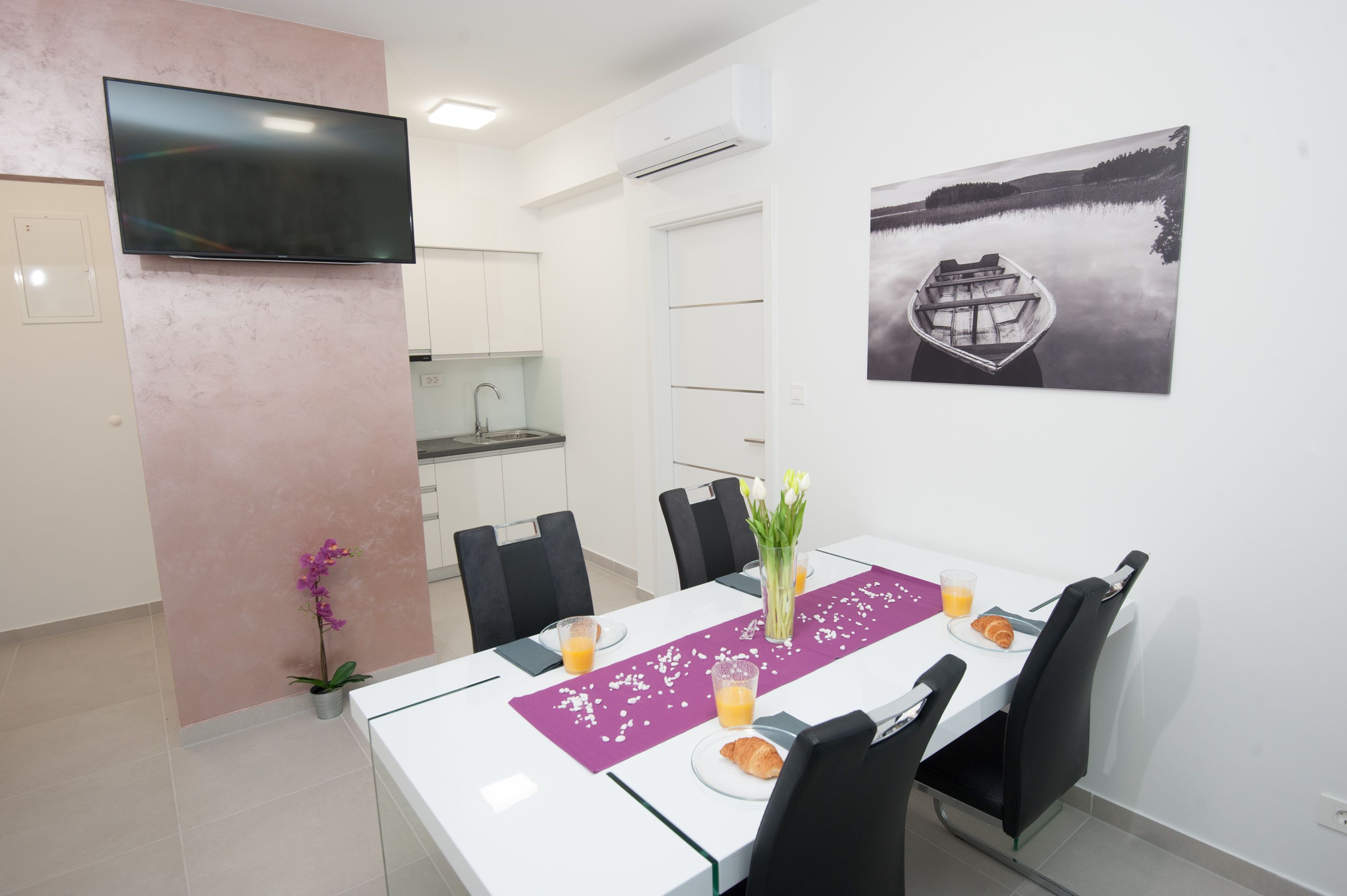 Uma sala de jantar com uma mesa branca, cadeiras pretas, um jogo americano roxo e uma fotografia a preto e branco na parede.