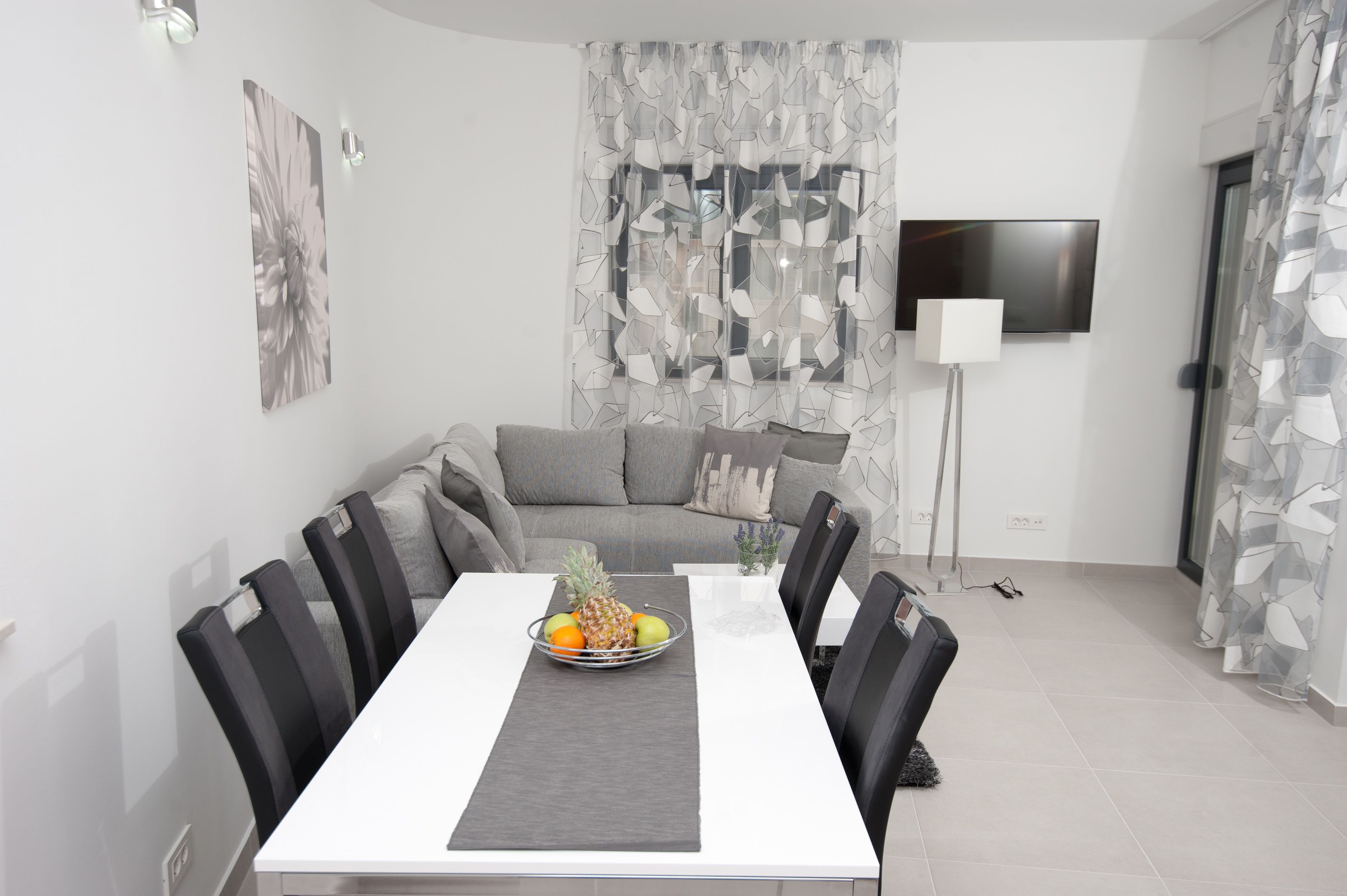 Ein moderner Essbereich mit einem weißen Tisch, schwarzen Stühlen und einer Obstschale. Im Hintergrund sind ein graues Sofa und ein Fernseher zu sehen.