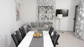 Ein moderner Essbereich mit einem weißen Tisch, schwarzen Stühlen und einer Obstschale. Im Hintergrund sind ein graues Sofa und ein Fernseher zu sehen.