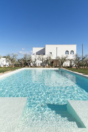 Una piscina rettangolare con acqua cristallina, circondata da un edificio bianco e alberi.