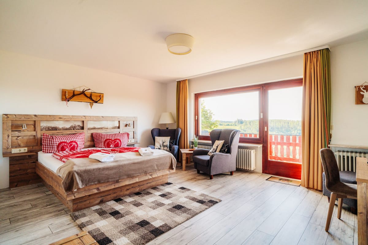 Ein Schlafzimmer mit einem hölzernen Bett, einem Schreibtisch, einem Stuhl und Blick ins Freie durch ein großes Fenster.