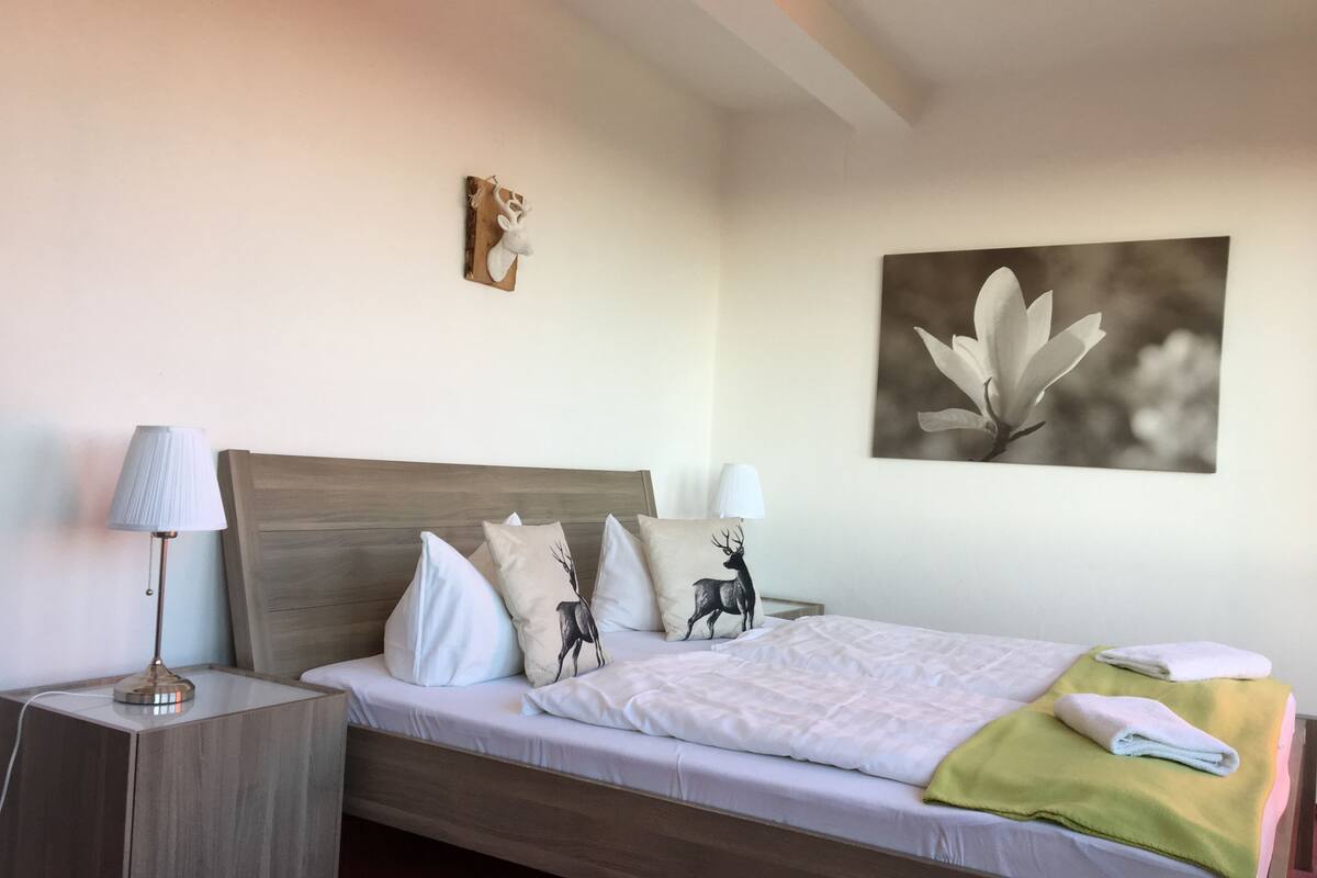 Ein ordentlich bezogenes Bett mit hölzernem Kopfteil, zwei Nachttischlampen und einem an der Wand hängenden Kunstwerk.