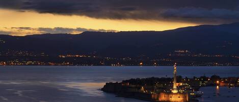 Un paisaje urbano costero al atardecer con un faro destacado en una isla, rodeado de agua y luces de ciudad a lo lejos.