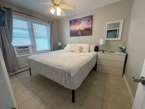 Ein Schlafzimmer mit einem Bett, einem Nachttisch, einem Deckenventilator, einem Fenster mit Jalousien, einem Wandbild und einer Tür.