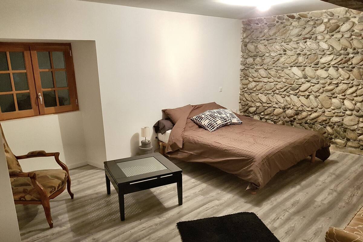 A bedroom with a bed, a chair, a table, and a stone wall.