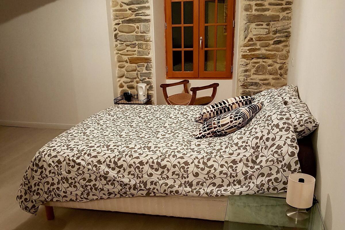 A bedroom with a bed, a chair, a table, a stone wall, and a wooden door.