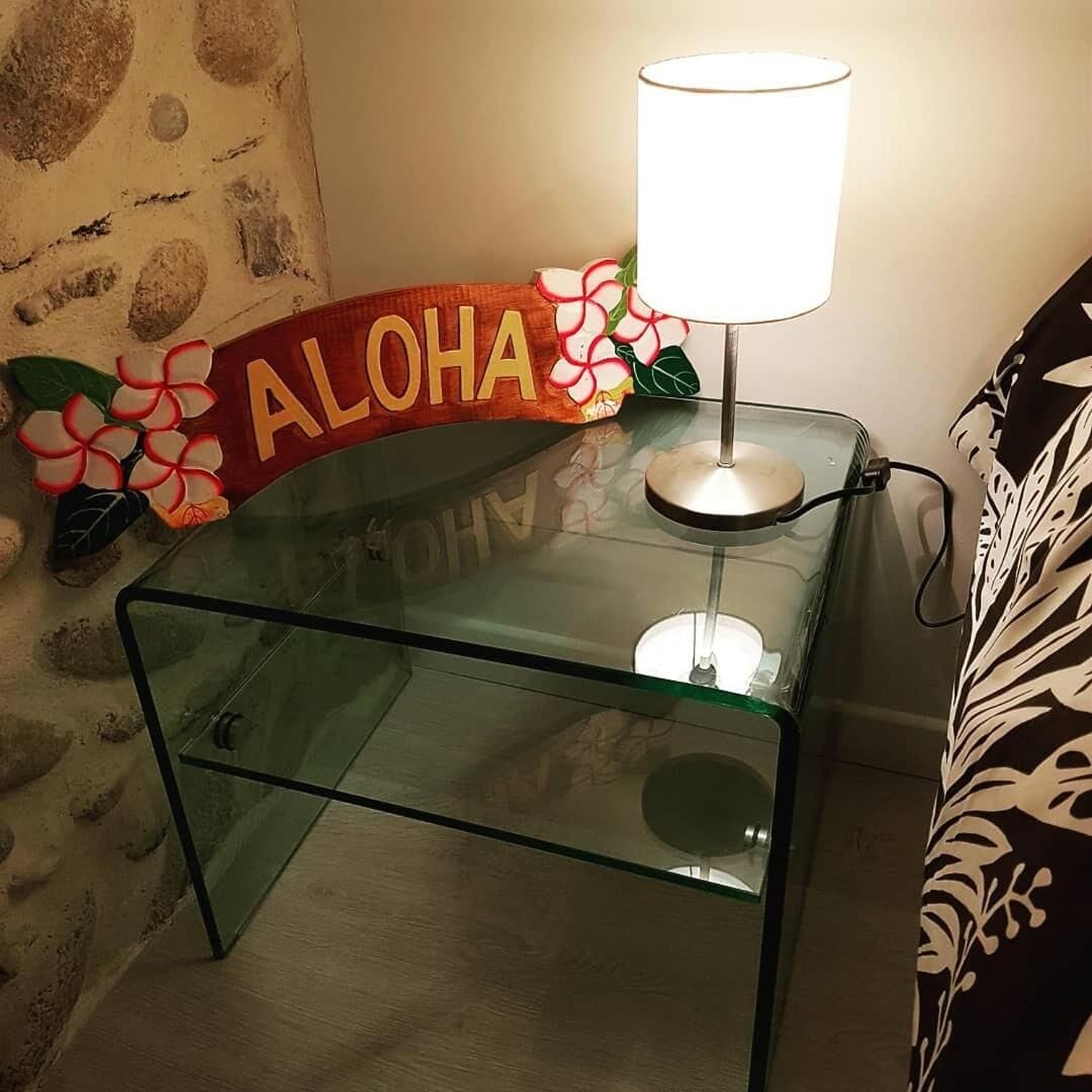 Ein Glastisch mit einer Lampe, einem Kissen mit 'ALOHA' und Blumen, sowie einer Steinsäule.