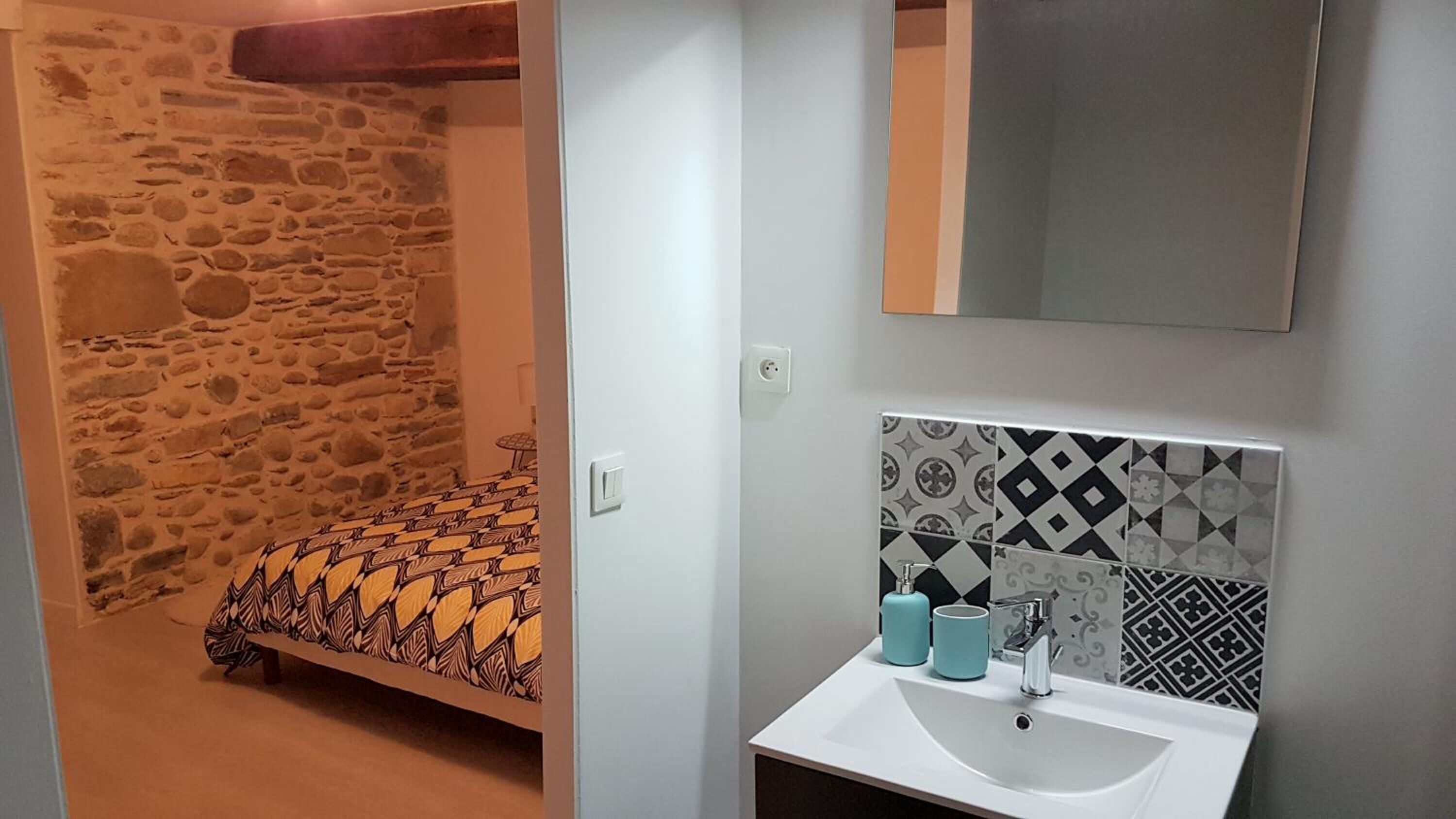 Une chambre avec un mur en pierre, une couvre-lit à motifs et une salle de bain avec des carreaux décoratifs et un lavabo.
