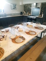 A dining table set with plates, bowls, and cutlery, a pitcher, and a pie in a kitchen with a stone backsplash and a gas stove.