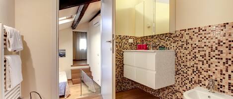 Casa de banho com lavatório branco, espelho e painel de azulejos. Uma porta dá acesso a um quarto com cama e escadas.