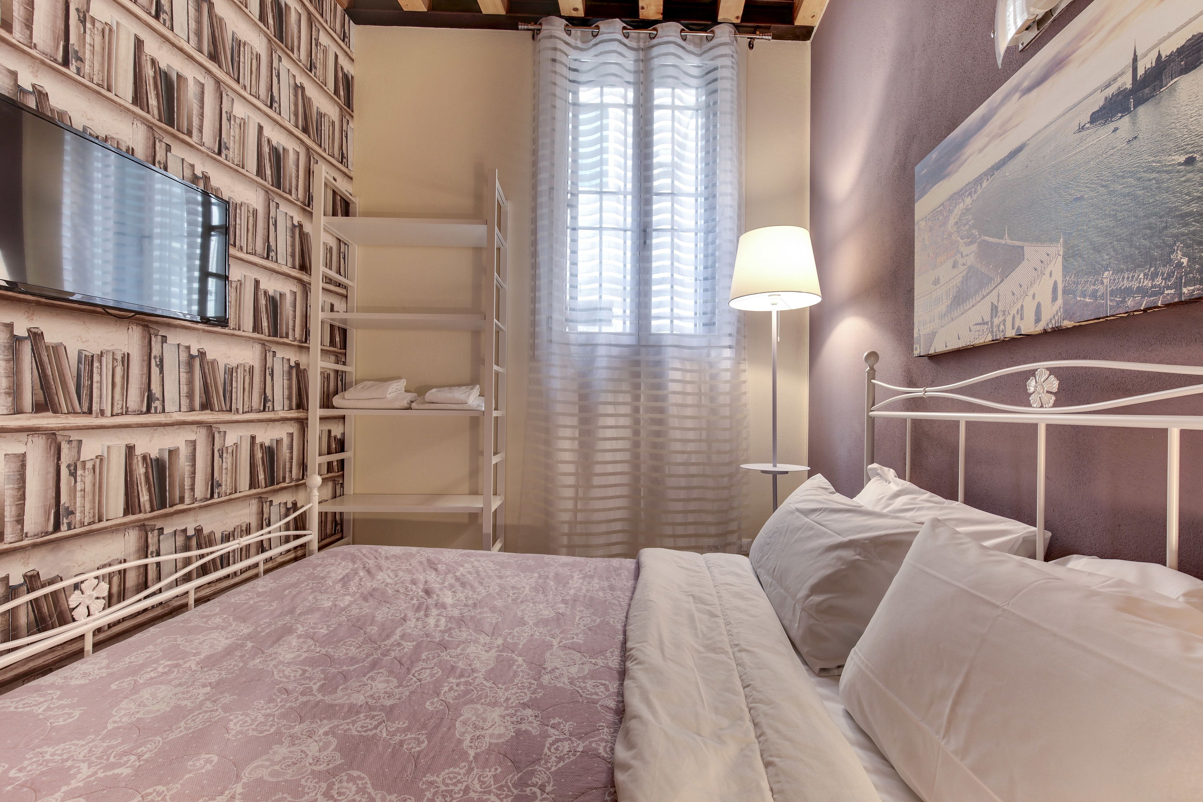 A bedroom with a bed, a bookshelf, a painting, and a window with curtains.